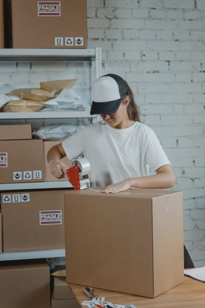pexels-photo-6169030 Young female worker sealing cardboard boxes in an indoor warehouse setting.